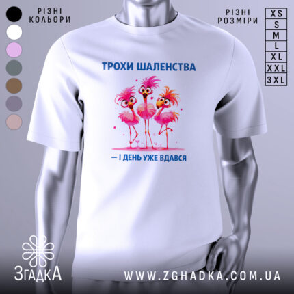 Футболка трохи шаленства, білого кольору, зображена з фронтального ракурсу на манекені, фон нейтральний, принт з трьома рожевими фламінго з веселими виразами обличчя та написом синього кольору.