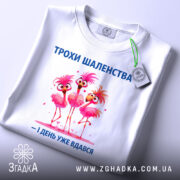 Футболка трохи шаленства, білого кольору, зображена з верхнього ракурсу, фон світлий, принт з трьома рожевими фламінго, напис синього кольору, на футболці є етикетка з зеленим шнурком.
