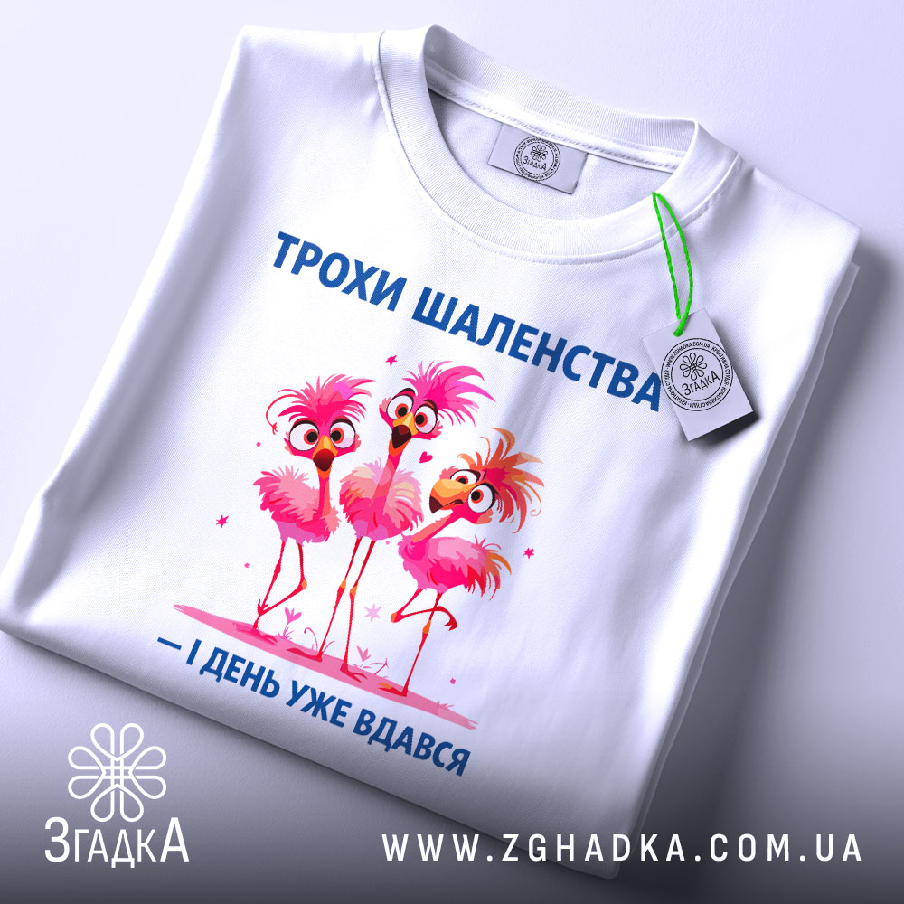 Футболка трохи шаленства, білого кольору, зображена з верхнього ракурсу, фон світлий, принт з трьома рожевими фламінго, напис синього кольору, на футболці є етикетка з зеленим шнурком.