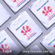 Футболка трохи шаленства, білого кольору, зображена з фронтального ракурсу на манекені, фон нейтральний, принт з трьома рожевими фламінго з веселими виразами обличчя та написом синього кольору.
