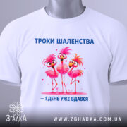Футболка трохи шаленства, білого кольору, зображена з верхнього ракурсу, фон світлий, принт з трьома рожевими фламінго, напис синього кольору, на футболці є етикетка з зеленим шнурком.