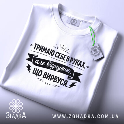 Футболка тримаю себе в руках, білого кольору, з верхнього ракурсу, складена на поверхні, фон світло-сірий, принт чорного кольору з текстом та етикеткою.