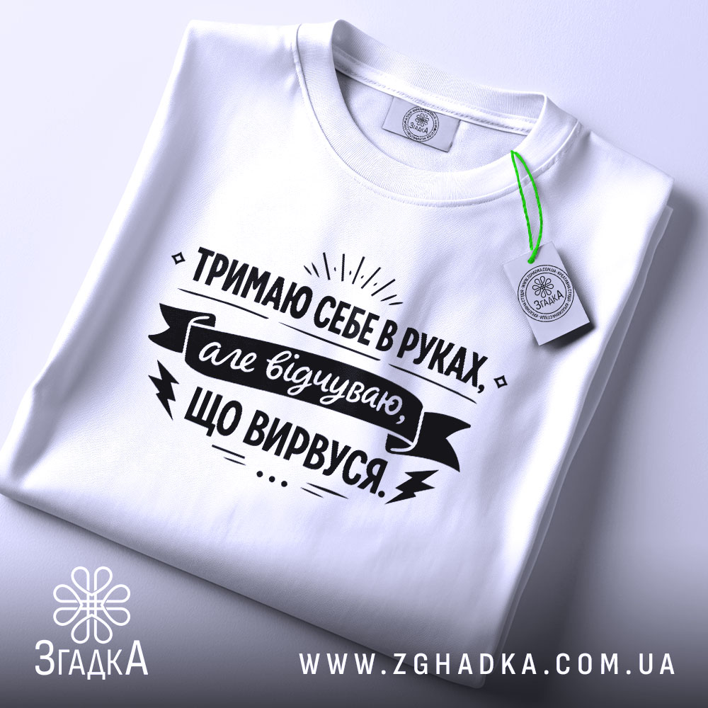 Футболка тримаю себе в руках, білого кольору, з верхнього ракурсу, складена на поверхні, фон світло-сірий, принт чорного кольору з текстом та етикеткою.