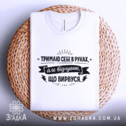 Футболка тримаю себе в руках, білого кольору, з верхнього ракурсу, складена на плетеній основі, фон світло-сірий, принт чорного кольору з текстом.