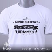 Футболка тримаю себе в руках, білого кольору, з верхнього ракурсу, складена на поверхні, фон світло-сірий, принт чорного кольору з текстом та етикеткою.