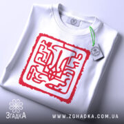 Футболка тризуб код нації, біла, складена на столі, фон світло-сірий, принт червоний з абстрактним тризубом, на комірці є етикетка з зеленим шнурком.