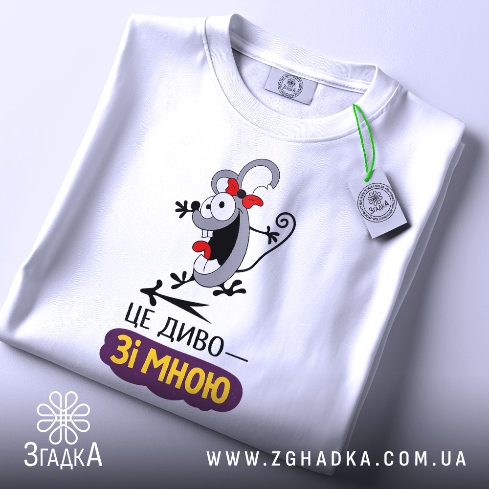 Футболка це диво. Біла футболка, складена на поверхні, з видимим принтом персонажа з червоною стрічкою. На футболці напис "ЦЕ ДИВО—ЗІ МНОЮ". На комірці видно етикетку, а фон однотонний, світлий.