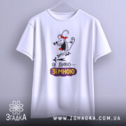 Футболка це диво. Біла футболка з коротким рукавом, підвішена на вішалці, з фронтального ракурсу. Принт з персонажем з червоною стрічкою та написом "ЦЕ ДИВО—ЗІ МНОЮ". Фон світлий, з легким градієнтом.