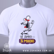 Футболка це диво. Біла футболка, складена на поверхні, з видимим принтом персонажа з червоною стрічкою. На футболці напис "ЦЕ ДИВО—ЗІ МНОЮ". На комірці видно етикетку, а фон однотонний, світлий.