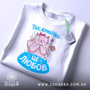 Футболка це любов. Біла футболка, складена на поверхні, з принтом кішки, яка обіймає пташку. Принт яскравий, з рожевими та блакитними елементами, а на футболці є етикетка з логотипом.