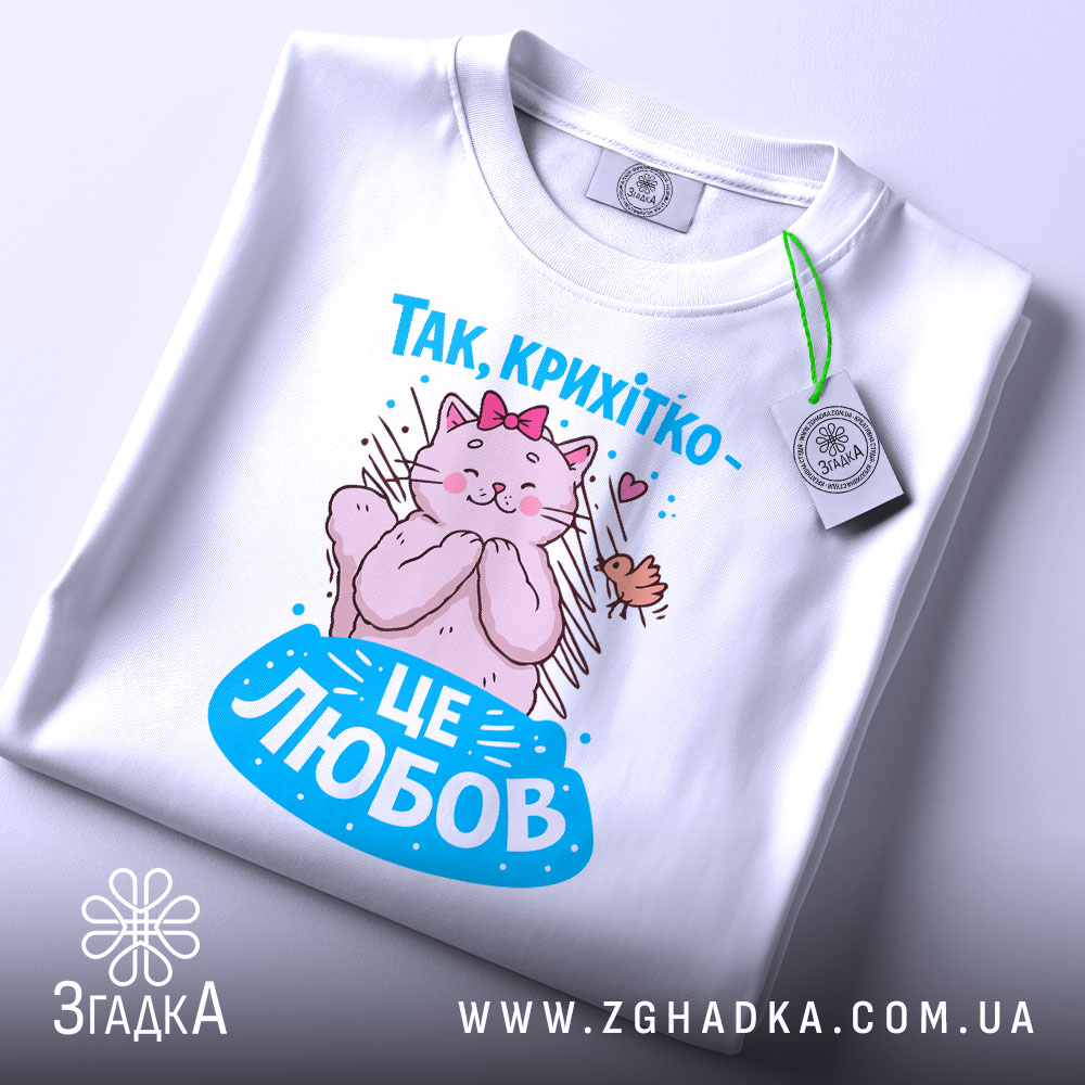 Футболка це любов. Біла футболка, складена на поверхні, з принтом кішки, яка обіймає пташку. Принт яскравий, з рожевими та блакитними елементами, а на футболці є етикетка з логотипом.
