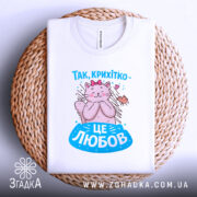 Футболка це любов. Біла футболка, складена на плетеній основі, з принтом кішки, що обіймає пташку. Принт яскравий, а фон створює затишну атмосферу, підкреслюючи стиль футболки.