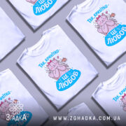 Футболка це любов. Біла футболка з принтом, на якому зображена кішка з рожевою стрічкою на голові, що обіймає пташку. Принт виконаний у яскравих кольорах, фон нейтральний, а футболка представлена на манекені.