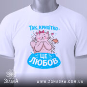 Футболка це любов. Біла футболка, складена на поверхні, з принтом кішки, яка обіймає пташку. Принт яскравий, з рожевими та блакитними елементами, а на футболці є етикетка з логотипом.