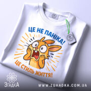 Футболка це не паніка. Біла футболка з принтом, на якому зображено переляканого кролика, що кричить. Принт яскравий, з жовтими та синіми елементами, на фоні світло-сірого кольору. Футболка зображена зверху, з етикеткою, що звисає.