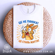 Футболка це не паніка. Біла футболка з принтом, на якому зображено переляканого кролика, що кричить. Принт яскравий, з жовтими та синіми елементами, на фоні світло-сірого кольору. Футболка лежить на плетеній кошику, з видимою етикеткою.