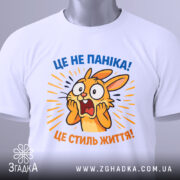 Футболка це не паніка. Біла футболка з принтом, на якому зображено переляканого кролика, що кричить. Принт яскравий, з жовтими та синіми елементами, на фоні світло-сірого кольору. Футболка зображена зверху, з етикеткою, що звисає.