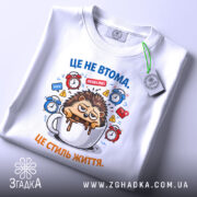 Футболка це не втома. Біла футболка, зображена зверху, з принтом їжачка у чашці, на фоні з годинниками, з етикеткою на комірці, ракурс зверху, м'яка текстура.