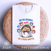 Футболка це не втома. Біла футболка, складена на плетеній основі, з принтом їжачка у чашці, ракурс зверху, фон натуральний, акцент на принті.