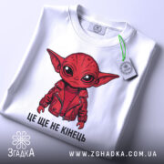 Футболка це ще не кінець. Біла футболка з принтом червоного персонажа, що одягнений у червону куртку, зображена з верхнього ракурсу на світлому фоні. На футболці видно етикетку з зеленим шнурком.