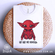 Футболка це ще не кінець. Біла футболка з принтом червоного персонажа в червоній куртці, зображена зверху на плетеній основі. Текст "ЦЕ ЩЕ НЕ КІНЕЦЬ" видно під принтом.
