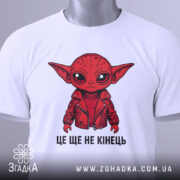 Футболка це ще не кінець. Біла футболка з принтом червоного персонажа, що одягнений у червону куртку, зображена з верхнього ракурсу на світлому фоні. На футболці видно етикетку з зеленим шнурком.