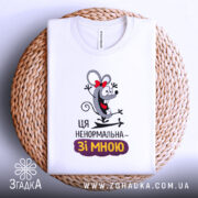 Футболка ця ненормальна. Білий колір, зображена зверху на плетеній основі, фон білий, принт з мишкою з червоною стрічкою, текст "Ця ненормальна – зі мною" під малюнком.