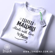 Футболка Циці Мациці білого кольору, складена на поверхні, фон світло-сірий, принт чорного кольору з написом 'Циці Мациці Made with love by Mom', на футболці є етикетка з зеленим шнурком.