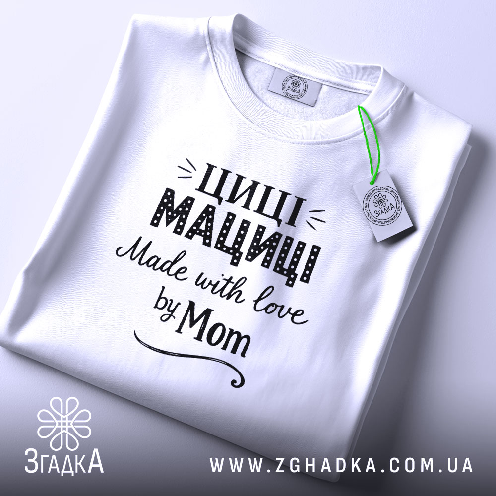 Футболка Циці Мациці білого кольору, складена на поверхні, фон світло-сірий, принт чорного кольору з написом 'Циці Мациці Made with love by Mom', на футболці є етикетка з зеленим шнурком.