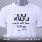 Футболка Циці Мациці білого кольору, складена на поверхні, фон світло-сірий, принт чорного кольору з написом 'Циці Мациці Made with love by Mom', на футболці є етикетка з зеленим шнурком.