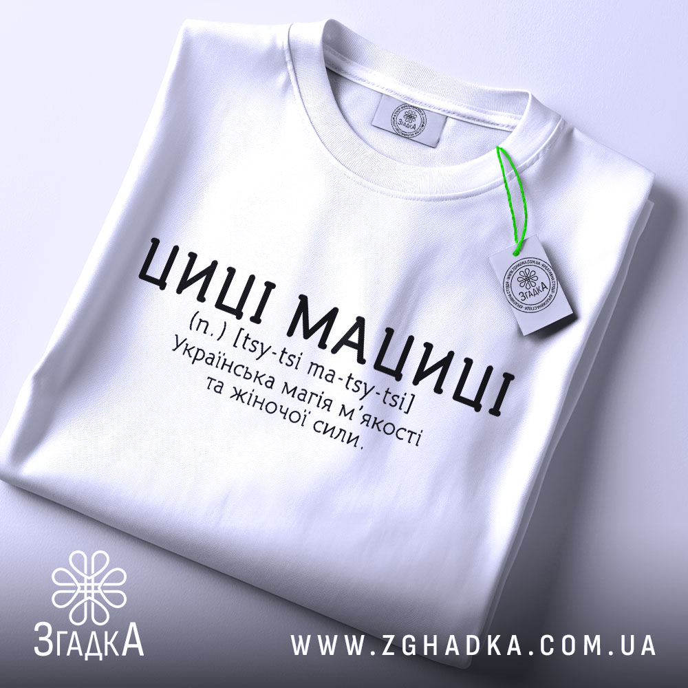 Футболка циці мациці білого кольору, складена на поверхні, фон світлий, принт чорного кольору з текстом про українську магію жіночої сили, на комірці видна етикетка.