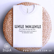 Футболка циці мациці білого кольору, складена на плетеній основі, фон світлий, принт чорного кольору з текстом про українську магію жіночої сили.