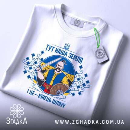 Футболка тут наша земля. Біла футболка, складена на поверхні, зображена з верхнього ракурсу. На фоні світло-сірий колір. Принт з чоловіком, що тримає меч, оточений декоративними елементами, а також зелена етикетка.