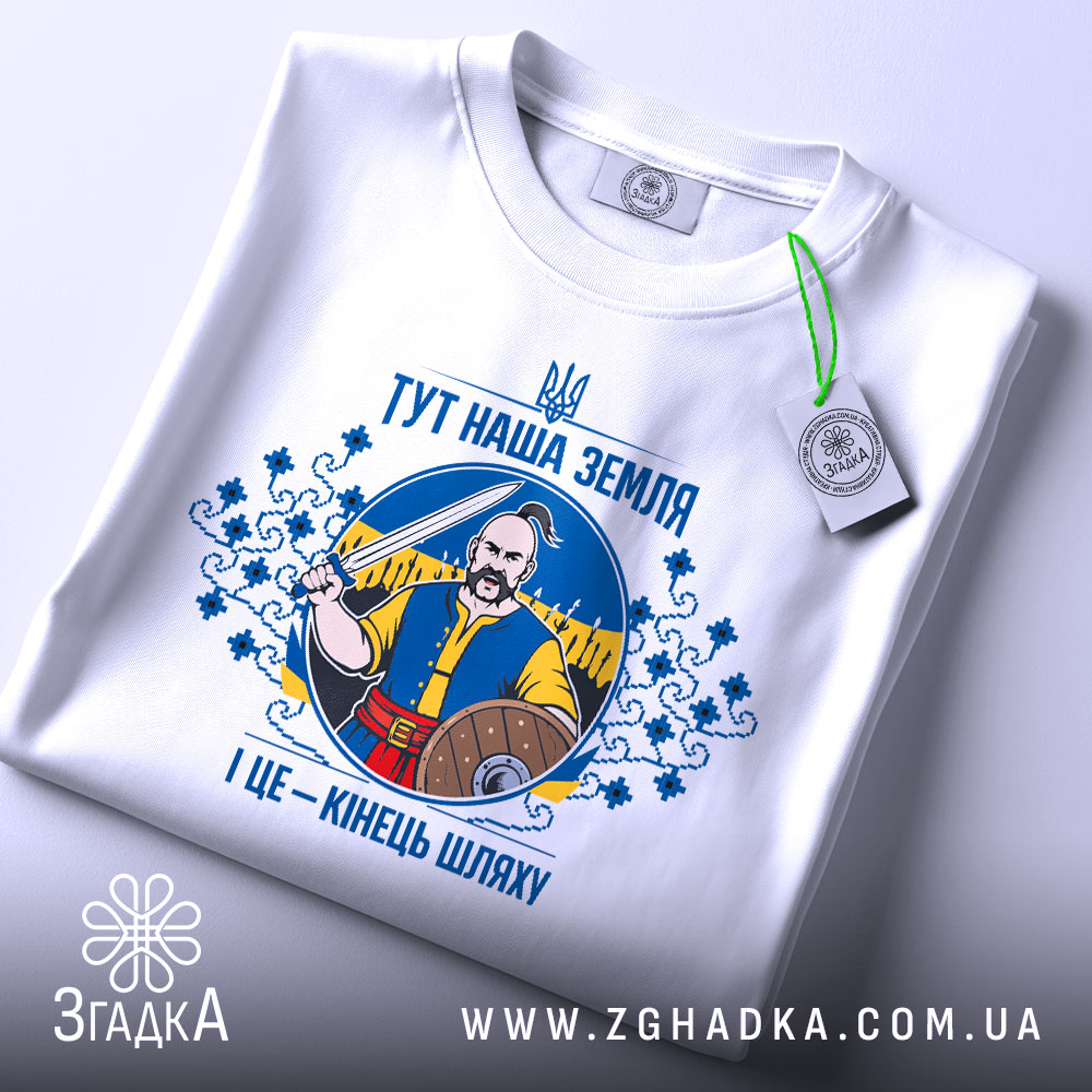 Футболка тут наша земля. Біла футболка, складена на поверхні, зображена з верхнього ракурсу. На фоні світло-сірий колір. Принт з чоловіком, що тримає меч, оточений декоративними елементами, а також зелена етикетка.