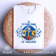 Футболка тут наша земля. Біла футболка, складена на плетеній основі, зображена з верхнього ракурсу. На фоні натуральний колір плетеного матеріалу. Принт з чоловіком, що тримає меч, оточений декоративними елементами у синьому та жовтому кольорах.