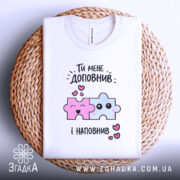 Футболка ти мене доповнив, біла, складена на плетеній кошику, фон білий, принт з двома кольоровими пазлами: рожевим і блакитним, з усмішками та сердечками.