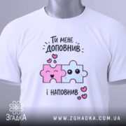 Футболка ти мене доповнив, біла, складена на поверхні, фон світло-сірий, принт з двома кольоровими пазлами: рожевим і блакитним, з усмішками та сердечками, з етикеткою на комірці.