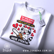 Футболка ти мені подобаєшся. Біла футболка, складена на поверхні, з принтом кота з бензопилою, видно етикетку, фон світлий, злегка текстурований.