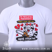 Футболка ти мені подобаєшся. Біла футболка, складена на поверхні, з принтом кота з бензопилою, видно етикетку, фон світлий, злегка текстурований.