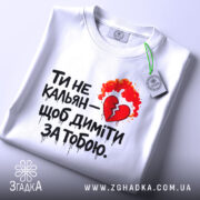 Футболка ти не кальян. Біла футболка, зображена зверху, на плоскій поверхні. На футболці великий принт з червоним серцем та вогнем. На комірці видно етикетку з логотипом бренду.