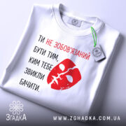 Футболка ти не зобов’язаний білого кольору, з принтом червоного кольору у вигляді маски та текстом, складена на поверхні, з етикеткою, фон світлий, без зайвих деталей.