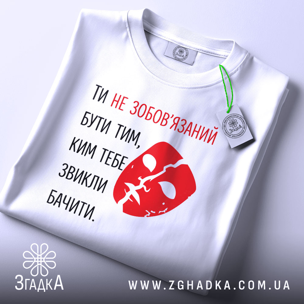 Футболка ти не зобов’язаний білого кольору, з принтом червоного кольору у вигляді маски та текстом, складена на поверхні, з етикеткою, фон світлий, без зайвих деталей.