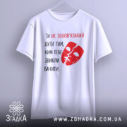 Футболка ти не зобов’язаний білого кольору, з принтом червоного кольору у вигляді маски та текстом, підвішена на вішалці, фон нейтральний, з легким градієнтом.