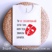 Футболка ти не зобов’язаний білого кольору, з принтом червоного кольору у вигляді маски та текстом, складена на плетеній основі, фон світлий, з натуральними текстурами.