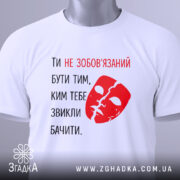 Футболка ти не зобов’язаний білого кольору, з принтом червоного кольору у вигляді маски та текстом, складена на поверхні, з етикеткою, фон світлий, без зайвих деталей.