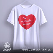 Футболка ти особлива людина. Біла футболка з червоним принтом у формі серця, на якому написано 'Ти особлива людина'. Футболка підвішена на вішалці з нейтральним фоном.