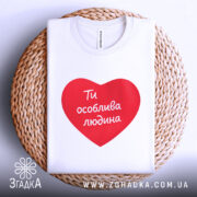 Футболка ти особлива людина. Біла футболка з червоним принтом у формі серця, на якому написано 'Ти особлива людина'. Футболка розташована на плетеній основі з нейтральним фоном.