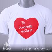 Футболка ти особлива людина. Біла футболка з червоним принтом у формі серця, на якому написано 'Ти особлива людина'. Футболка складена на білому фоні, з видимим етикеткою.
