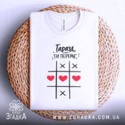 Футболка ти переміг, складена біла футболка на плетеній основі, фон світлий, принт з чорним текстом і червоними сердечками, показана зверху.