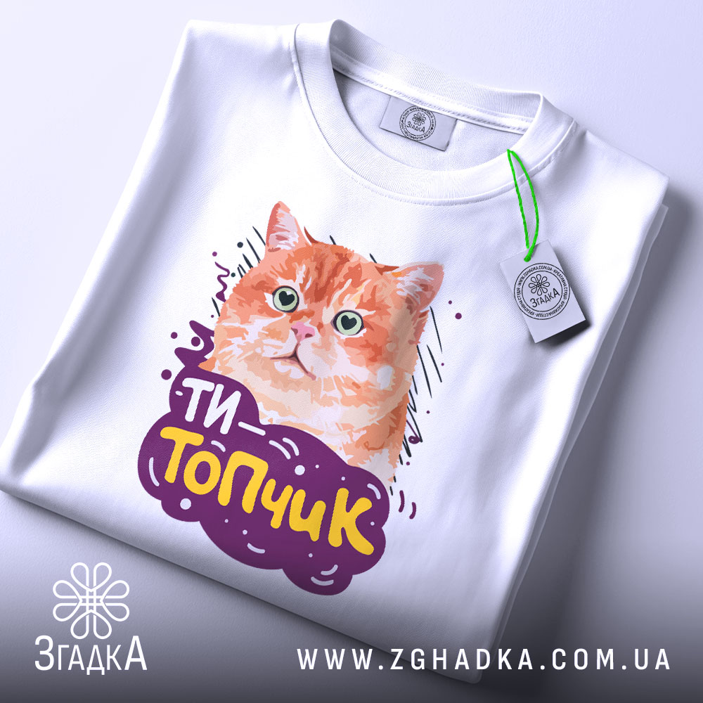 Футболка ти топчик, біла, зображена зверху, фон світло-сірий, принт з оранжевим котом та написом 'ТИ-Топчик', на футболці є етикетка з зеленим шнурком.